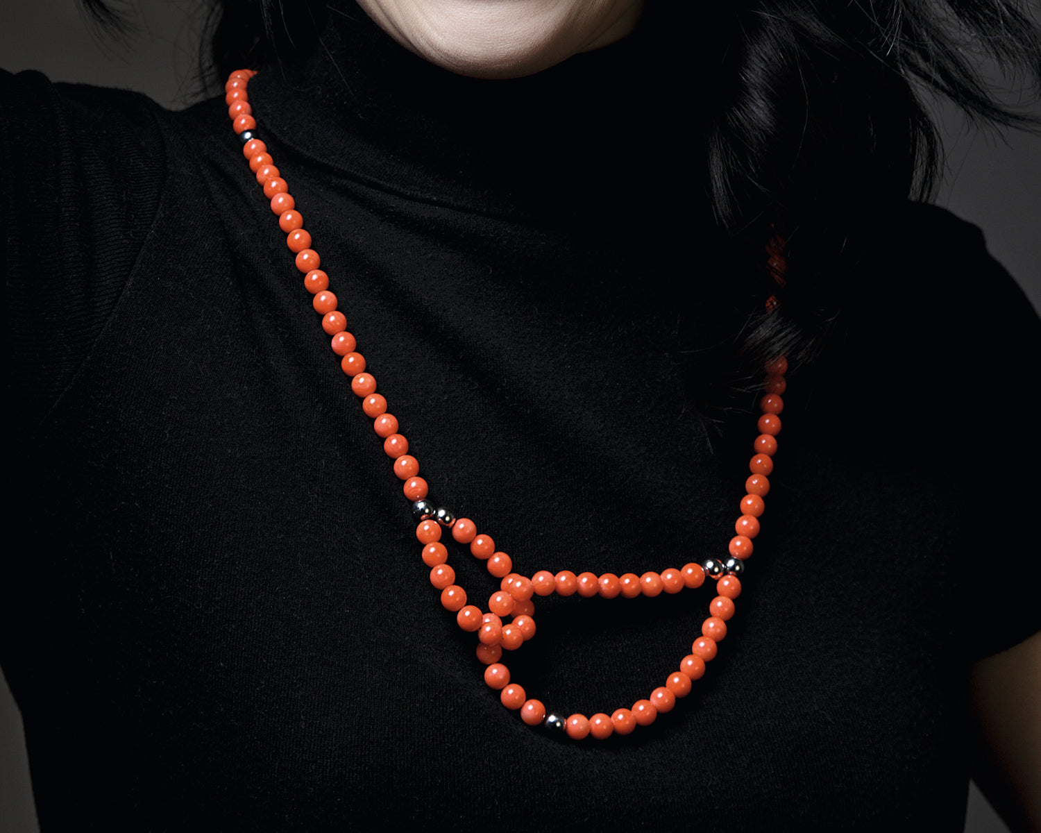 Zu sehen ist das Dekolleté einer Frau mit einer Kette aus orangeroten Bambuskorallekugeln auf einem schwarzen Oberteil. Ein Kettenende ist als kleine Schlaufe geformt, durch die das andere Kettenende geschlungen ist und als größere Schlaufe an der restlichen Kette angedockt ist. Dies ist eine der vielen Tragevarianten dieser wandelbaren Kette.
