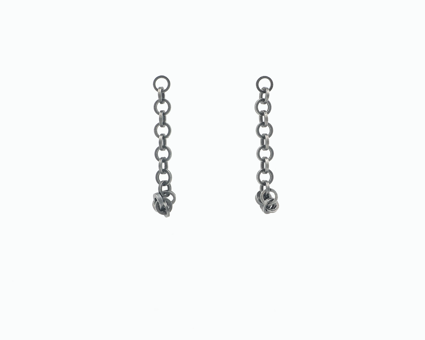 Zu sehen ist hier ein hängendes Ohrschmuck-Paar. Es sind zwei mit der Vorderseite nach vorne zeigende und nach unten hängende Kettenstücke aus ineinander eingehängten Kreisringelementen mit Vierkantprofil. Am unteren Ende sind ein paar Kettenelemente jeweils an den unten sitzenden Magneten angedockt. Dies ist eine der vielen Tragemöglichkeiten dieses Ohrschmuckes. Der Ohrschmuck ist anthrazitfarben. Er ist aus Edelstahl, Ruthenium geschwärzt.