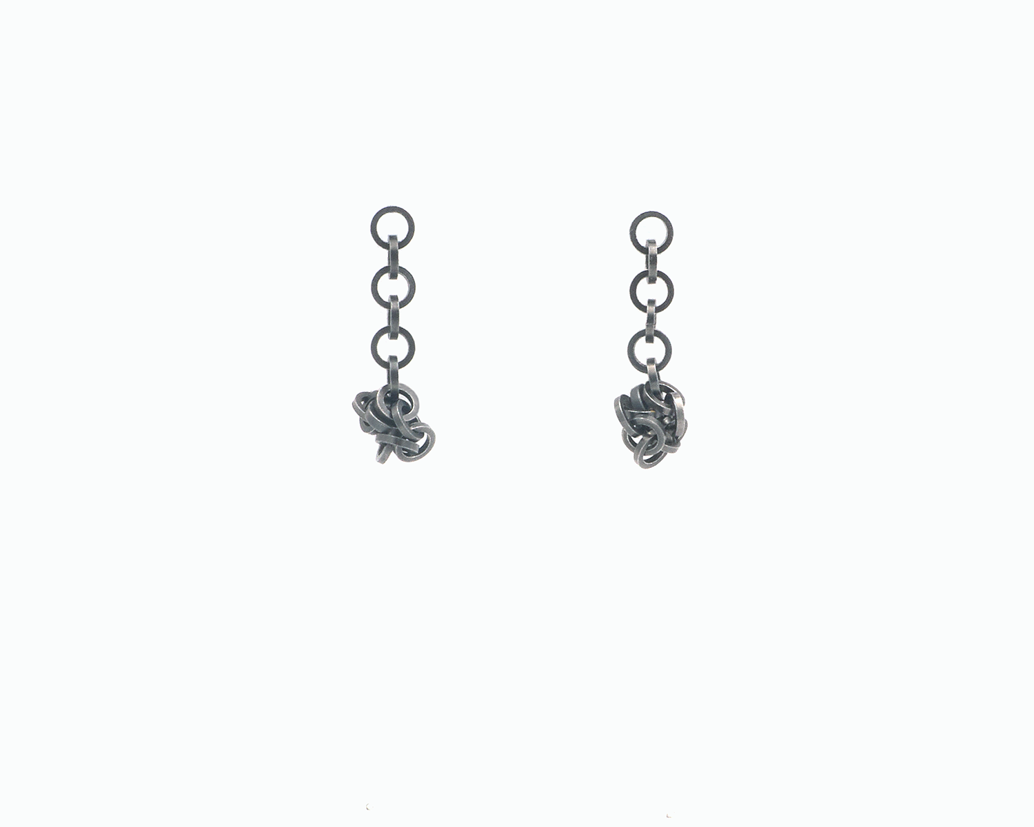 Zu sehen ist hier ein hängendes Ohrschmuck-Paar. Es sind zwei mit der Vorderseite nach vorne zeigende und nach unten hängende Kettenstücke aus ineinander eingehängten Kreisringelementen mit Vierkantprofil. Ungefähr die Hälfte des Kettenstranges ist jeweils an den unten sitzenden Magneten angedockt. Dies ist eine der vielen Tragemöglichkeiten dieses Ohrschmuckes. Der Ohrschmuck ist anthrazitfarben. Er ist aus Edelstahl, Ruthenium geschwärzt.