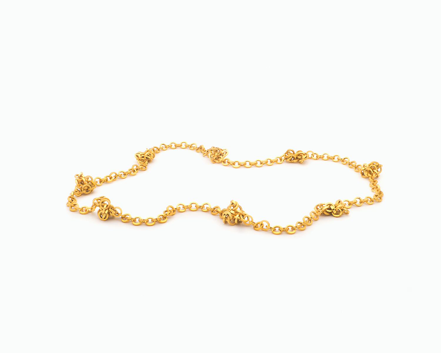 Zu sehen ist hier eine Kette aus runden, vierkantigen Ring-Elementen. Die Kette ist hier mit vereinzelten kleinen vermeintlichen Knotenpunkten dargestellt. An diesen“Knotenpunkten“ tummeln sich ein paar Ringelemente der Kette, da sie dort von in der Kette eingearbeiteten Magneten angezogen werden. Dies ist eine der vielen Tragevarianten dieser wandelbaren Kette. Die gesamte Kette ist goldfarben (Feingold plattiert).