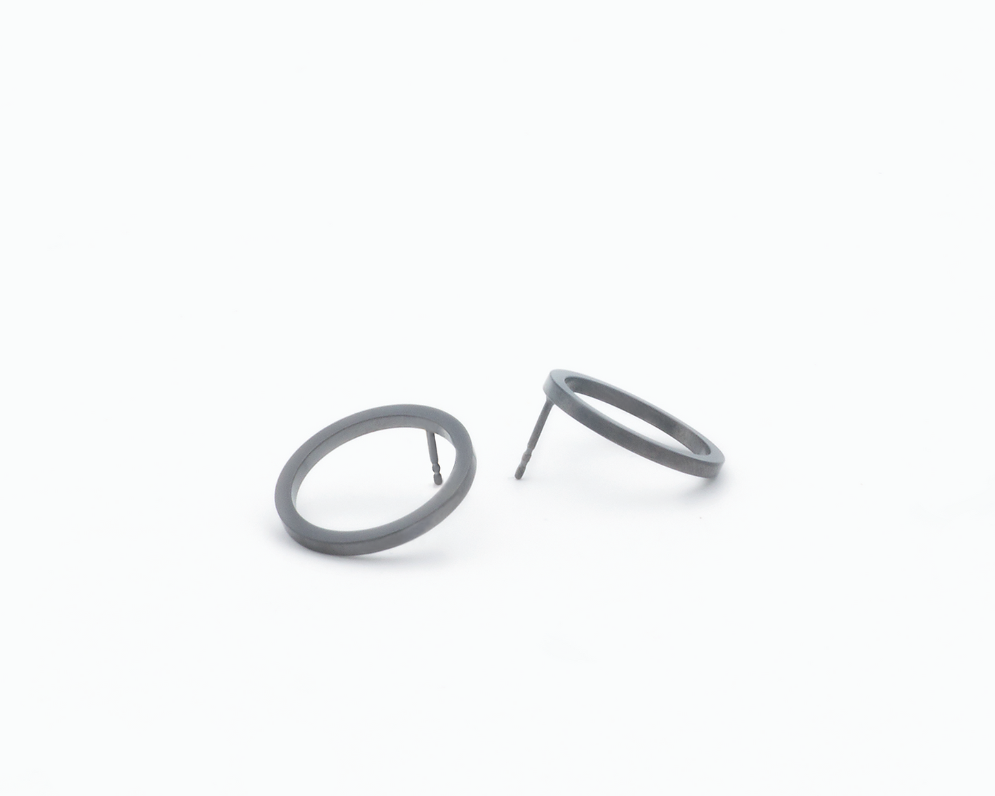 Zu sehen ist hier ein Ohrschmuck-Paar. Es sind zwei in unterschiedlicher Perspektive liegende, circa münzgroße, kreisförmige Ringe mit Vierkantprofil, an deren Rückseiten jeweils der Steckerstift sitzt. Der Ohrschmuck ist anthrazitfarben. Er ist aus Edelstahl, Ruthenium geschwärzt.
