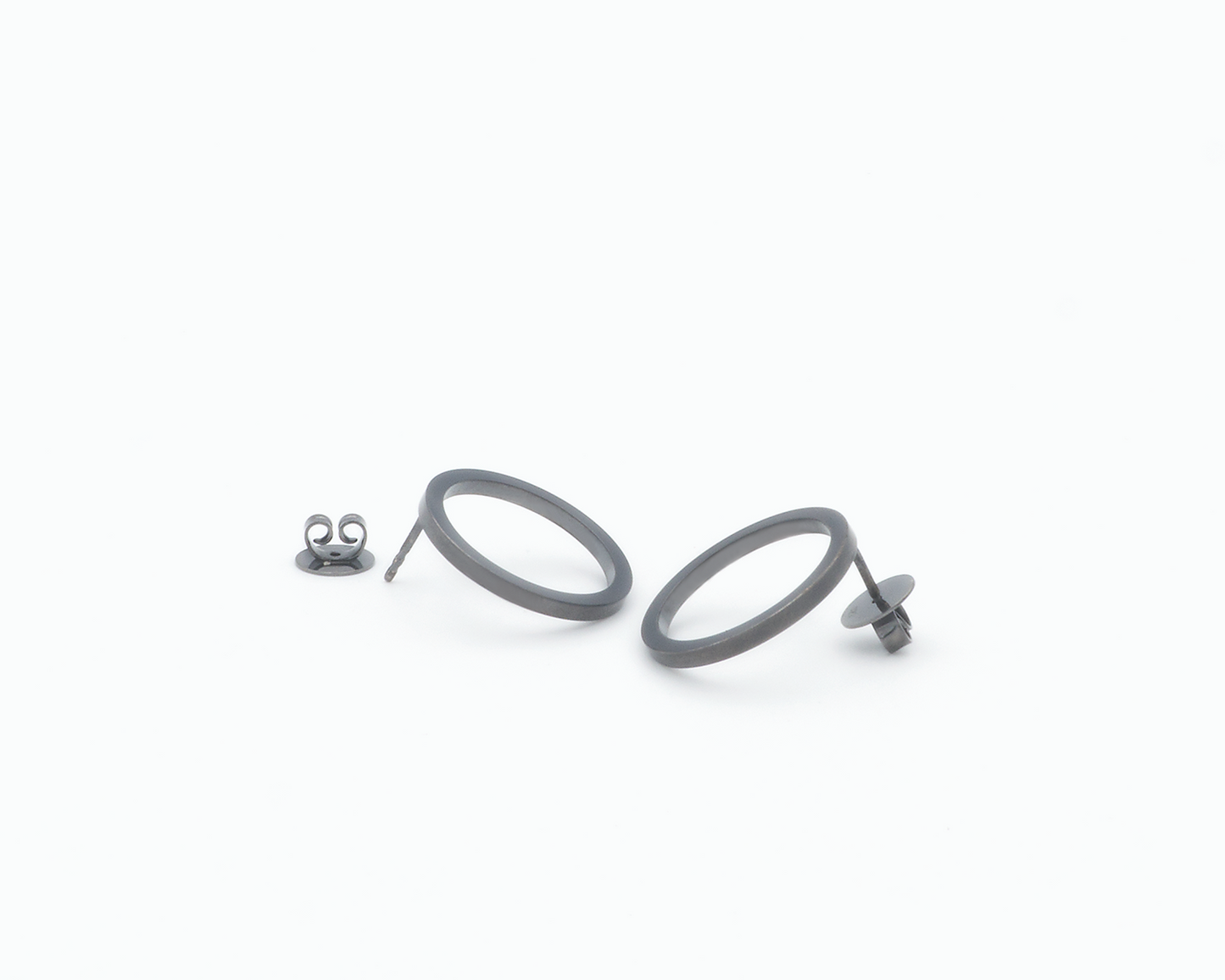 Zu sehen ist hier ein Ohrschmuck-Paar. Es sind zwei in unterschiedlicher Perspektive liegende, circa münzgroße, kreisförmige Ringe mit Vierkantprofil, an deren Rückseiten jeweils der Steckerstift sitzt. Beim linken Ohrschmuck liegt der Rückstecker daneben, beim rechten Ohrring steckt er auf dem Stift. Der Ohrschmuck ist anthrazitfarben. Er ist aus Edelstahl, Ruthenium geschwärzt.