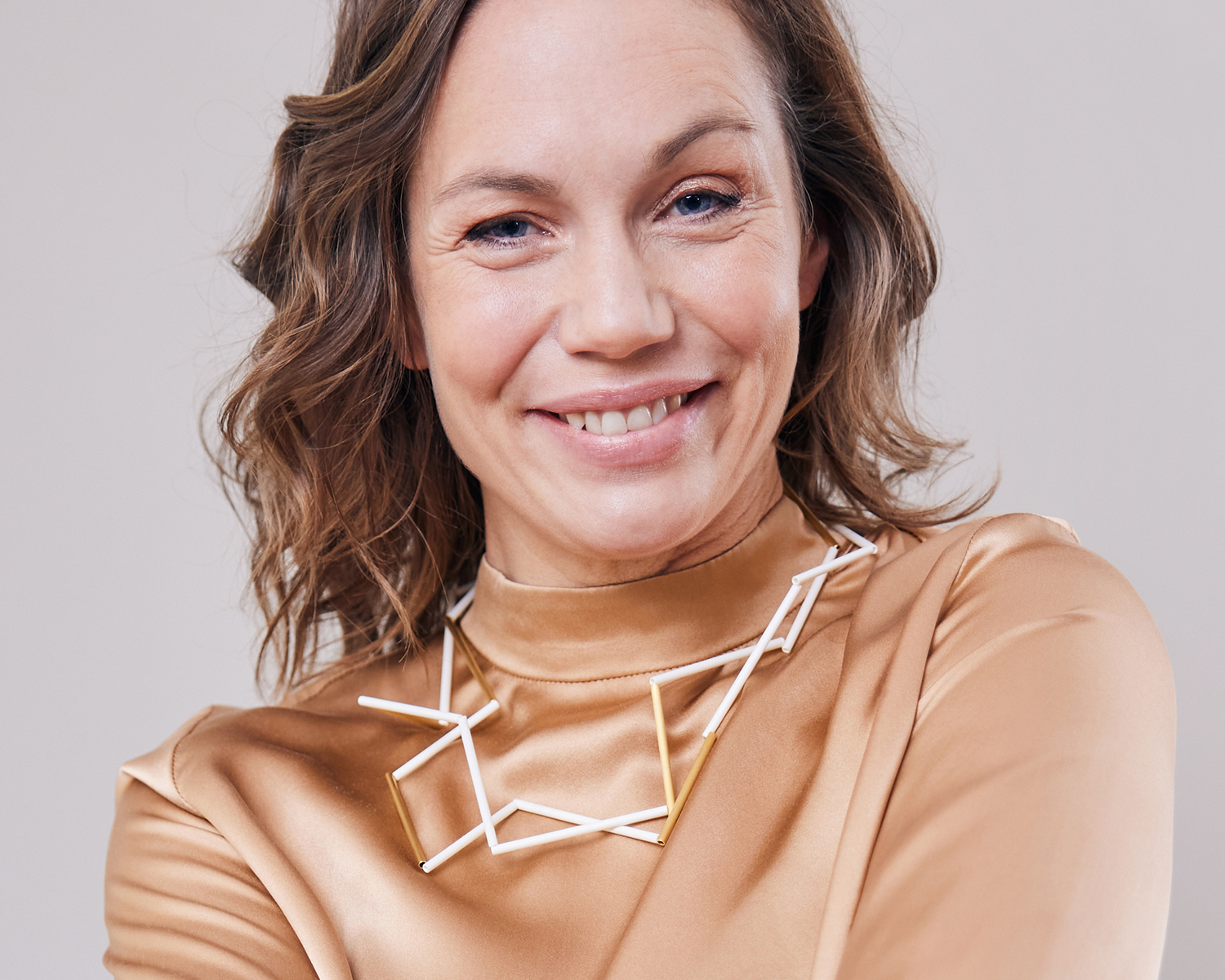 Zu sehen ist hier eine Frau mit beige glänzendem Oberteil, die eine Kette aus weißen und goldfarbenen Stäben trägt. Die Stäbe sind Rohrabschnitte in zwei verschiedenen Längen. Die Kette ist doppelt gelegt dargestellt und stellt somit eine kurze Tragevariante dar. Die Kette hat eine grafische Anmutung, bei der die Linie als Hauptakteur dominiert.