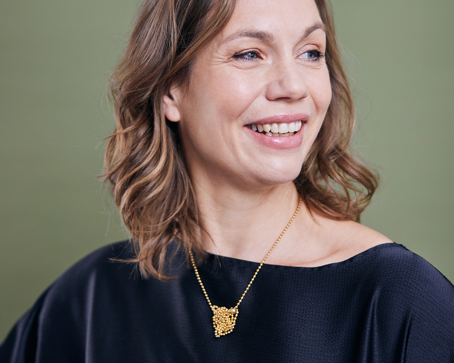 Zu sehen ist hier eine lächelnde, zur Seite blickende Frau mit schwarzem Oberteil, die eine Kugelkette mit weiteren, vereinzelt integrierten größeren Kugeln trägt. Vorne am Dekolleté sitzt ein scheinbarer Kettenknäuel (der allerdings durch einfaches Ziehen wieder aufgelöst werden kann).Der gesamte Halsschmuck ist goldfarben (Feingold plattiert).