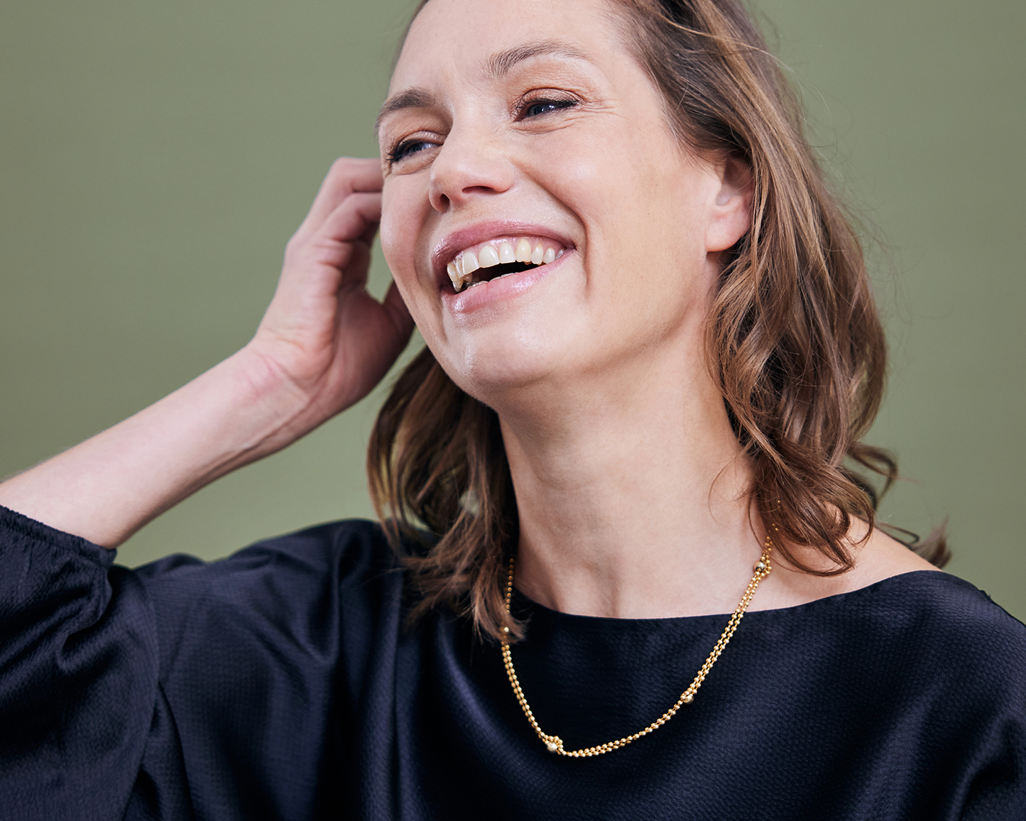 Zu sehen ist hier eine fröhlich lachende Frau mit schwarzem Oberteil, die eine Kugelkette mit weiteren, vereinzelt integrierten größeren Kugeln trägt. Die Kette hängt doppelt um den Hals. Der gesamte Halsschmuck ist goldfarben (Feingold plattiert).