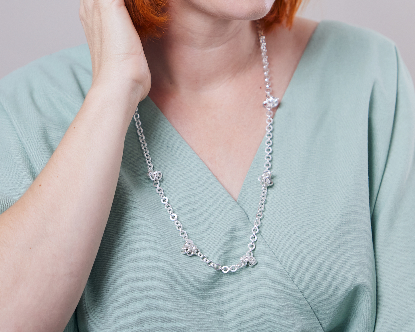 Zu sehen ist hier das Dekolleté einer Frau mit zartgrünem Oberteil, die eine Kette aus runden, vierkantigen Ring-Elementen trägt. Die Kette wird mittellang getragen mit vereinzelt sitzenden kleinen vermeintlichen Knotenpunkten. An diesen“Knotenpunkten“ tummeln sich ein paar Ringelemente der Kette, da sie dort von in der Kette eingearbeiteten Magneten angezogen werden. Dies ist dank der Wandelbarkeit der Kette eine der vielen Trageoptionen. Die gesamte Kette ist silberfarben (Silber plattiert).