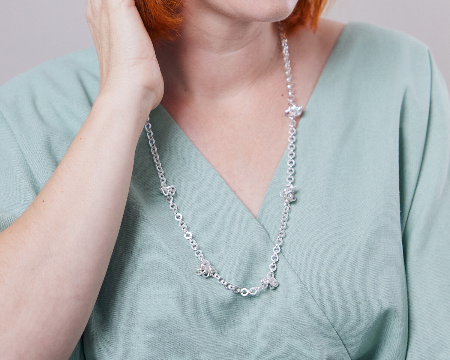 Zu sehen ist hier das Dekolleté einer Frau mit zartgrünem Oberteil, die eine Kette aus runden, vierkantigen Ring-Elementen trägt. Die Kette wird mittellang getragen mit vereinzelt sitzenden kleinen vermeintlichen Knotenpunkten. An diesen“Knotenpunkten“ tummeln sich ein paar Ringelemente der Kette, da sie dort von in der Kette eingearbeiteten Magneten angezogen werden. Dies ist dank der Wandelbarkeit der Kette eine der vielen Trageoptionen. Die gesamte Kette ist silberfarben (Silber plattiert).