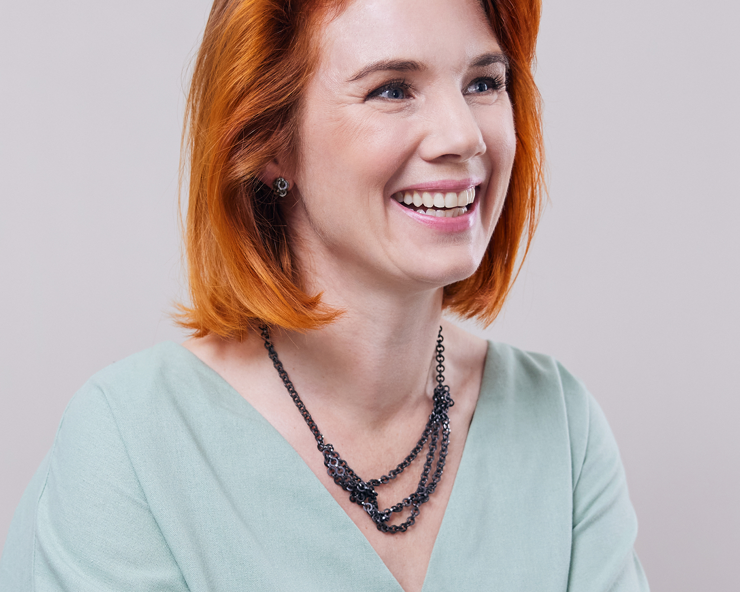 Zu sehen ist hier der Kopf und das Dekolleté einer Frau mit zartgrünem Oberteil, die eine Kette aus runden, vierkantigen Ring-Elementen trägt. Die Kette wird kurz getragen und hängt am Dekolleté dreireihig mit seitlich dickeren Abschnitten. Dies ist dank der Wandelbarkeit der Kette eine der vielen Trageoptionen. Die gesamte Kette ist dunkel, anthrazitfarben (Ruthenium geschwärzt).