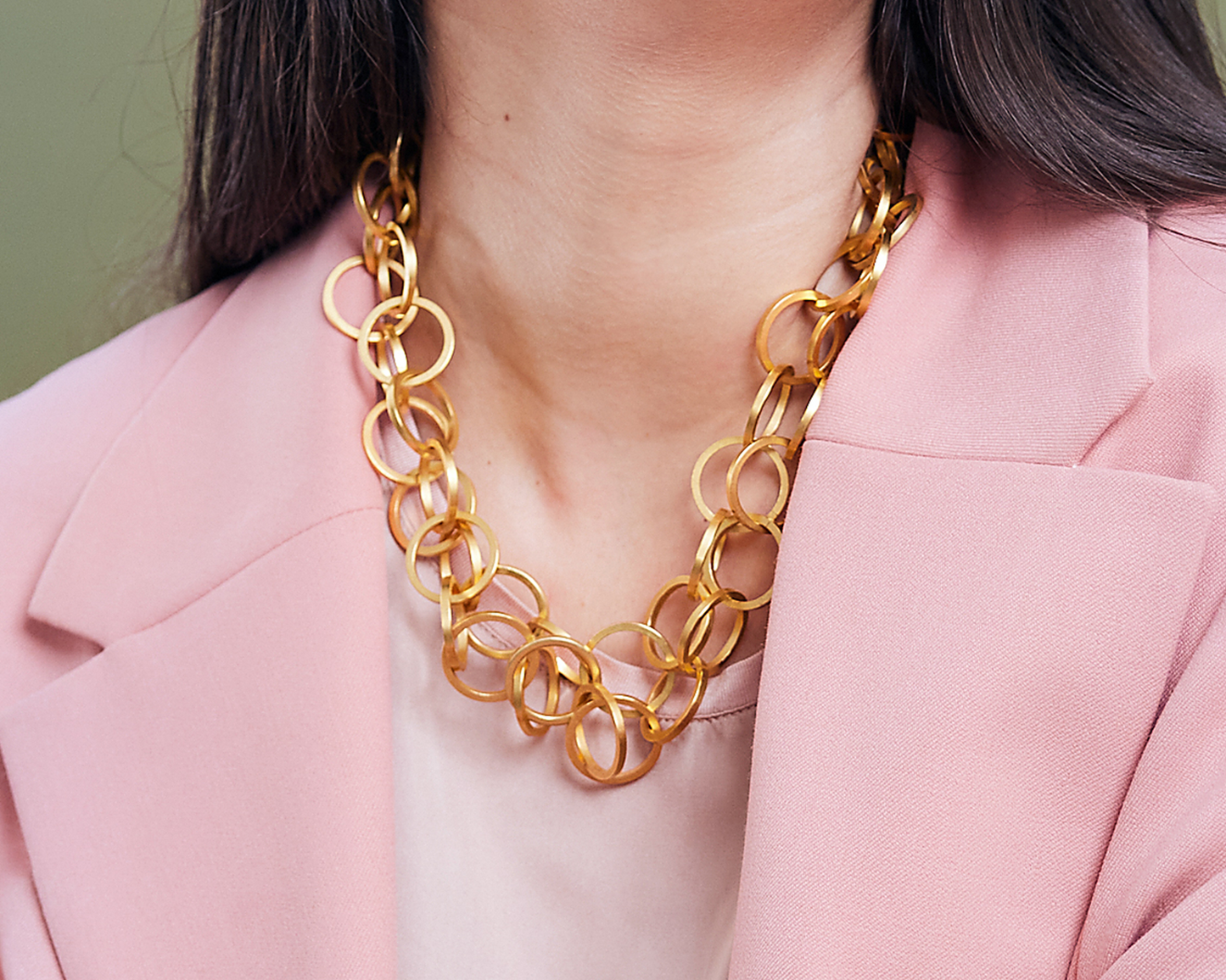 Zu sehen ist hier eine Kette aus runden, circa münzgroßen Ring-Elementen mit einem Vierkantprofil, getragen von einer Frau mit rosafarbenem Blazer. Auf dem Bild sieht man nur den Ausschnitts des Dekolletés. Die Ringe/Kettenglieder sind jeweils durch Einhängen mit dem nächsten Ring verbunden. Die Kette ist hier doppelt um den Hals gelegt, so dass sie wie eine kurze Kette aussieht. Dies ist eine der vielen Trageoptionen dieser Kette. Alle Ringe sind goldfarben (Feingold plattiert).