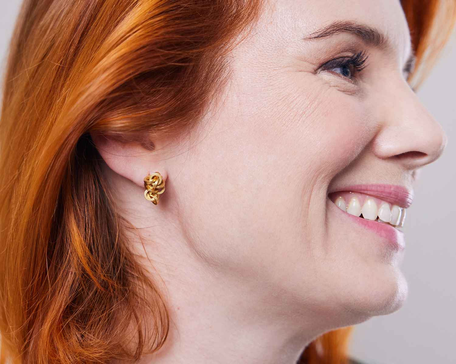 Zu sehen ist hier der zur Seite zeigende Kopf einer Frau, die den Ohrschmuck „attraente“ in Feingold plattierter Version trägt. In dem sichtbaren Ohr wird der Ohrstecker als „Kettenknäuel“ geformt, also als kurze Tragevariante gezeigt. Dies ist eine der vielen Trageoptionen dieses Ohrschmuckes.