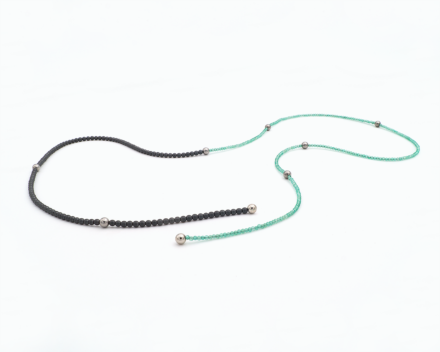 Zu sehen ist hier eine Kette aus etwas größeren dunkelgrauen, matten Hämatitkugeln und etwas kleineren grünen, facettierten Achatkugeln. Dazwischen sitzen Edelstahlkugeln und an den Enden Magnetkugeln. Die Kette ist offen und leicht kurvig liegend dargestellt. Dies ist eine der vielen Tragevarianten dieser wandelbaren Kette.