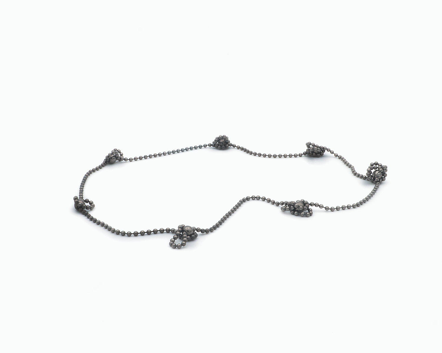 Zu sehen ist hier eine Kugelkette mit weiteren, vereinzelt integrierten größeren Kugeln. Die Kette ist hier mit vereinzelten kleinen vermeintlichen Knotenpunkten dargestellt. An diesen “Knotenpunkten“ tummeln sich Kettenabschnitte, da sie dort von den in der Kette eingearbeiteten Magnetkugeln angezogen werden. Dies ist eine der vielen Tragevarianten dieser wandelbaren Kette. Die gesamte Kette ist dunkel, anthrazitfarben (Ruthenium geschwärzt).