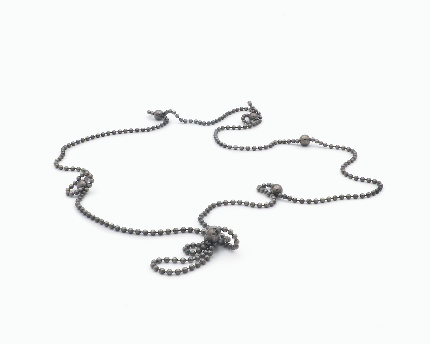 Zu sehen ist hier eine Kugelkette mit weiteren, vereinzelt integrierten größeren Kugeln. Die Kette ist hier in ihrer ganzen Länge  liegend und auf kurvige Weise präsentiert.  An manchen Stellen haben sich Kettenabschnitte an die nächstliegende Magnetkugel angedockt und bilden dort kleine Schlaufen. Die gesamte Kette ist dunkel, anthrazitfarben (Ruthenium geschwärzt).