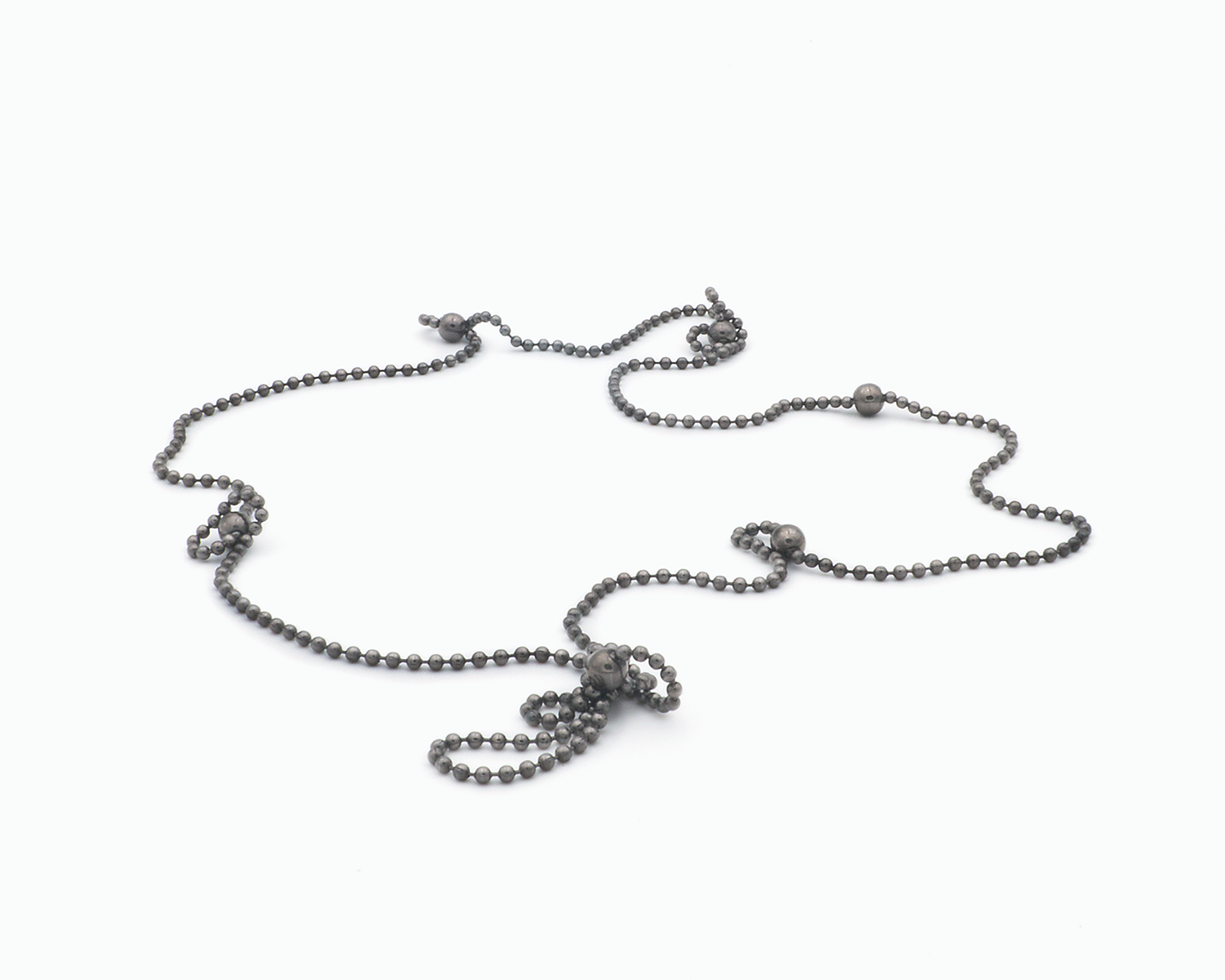 Zu sehen ist hier eine Kugelkette mit weiteren, vereinzelt integrierten größeren Kugeln. Die Kette ist hier in ihrer ganzen Länge  liegend und auf kurvige Weise präsentiert.  An manchen Stellen haben sich Kettenabschnitte an die nächstliegende Magnetkugel angedockt und bilden dort kleine Schlaufen. Die gesamte Kette ist dunkel, anthrazitfarben (Ruthenium geschwärzt).