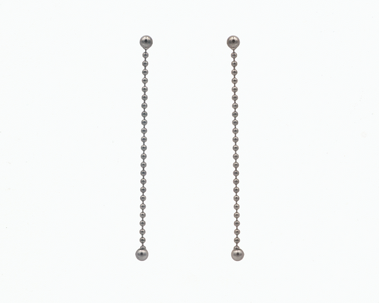 Zu sehen ist hier ein hängendes Ohrschmuck-Paar. Es sind zwei mit der Vorderseite nach vorne zeigende und nach unten hängende Kugelkettenstücke, an deren Abschluss unten jeweils eine größere Magnetkugel sitzt. Der Ohrschmuck ist anthrazitfarben. Er ist aus Edelstahl, Ruthenium geschwärzt.