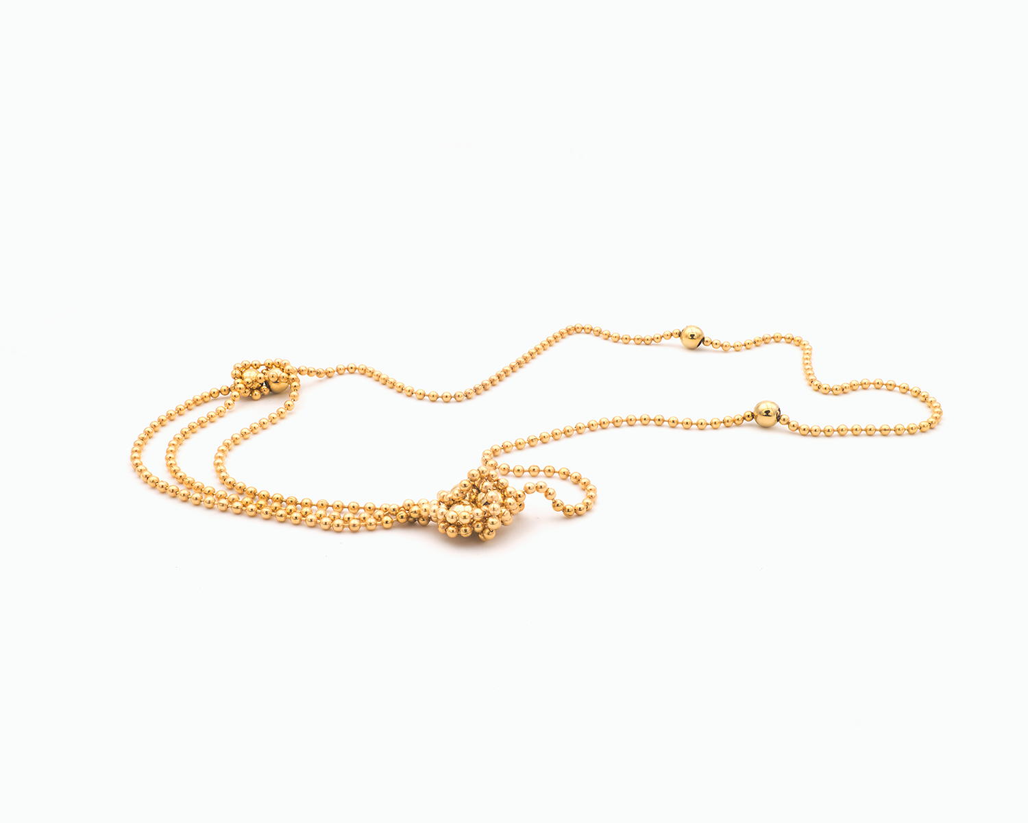 Zu sehen ist hier eine Kugelkette mit weiteren, vereinzelt integrierten größeren Kugeln. Die Kette ist liegend in einer Tragevariante dargestellt, bei der die Kette vorne im Dekolletébereich dreireihig liegt mit seitlich dickeren Abschnitten. Dies ist eine der vielen Trageoptionen dieser wandelbaren Kette. Die gesamte Kette ist goldfarben (Feingold plattiert).