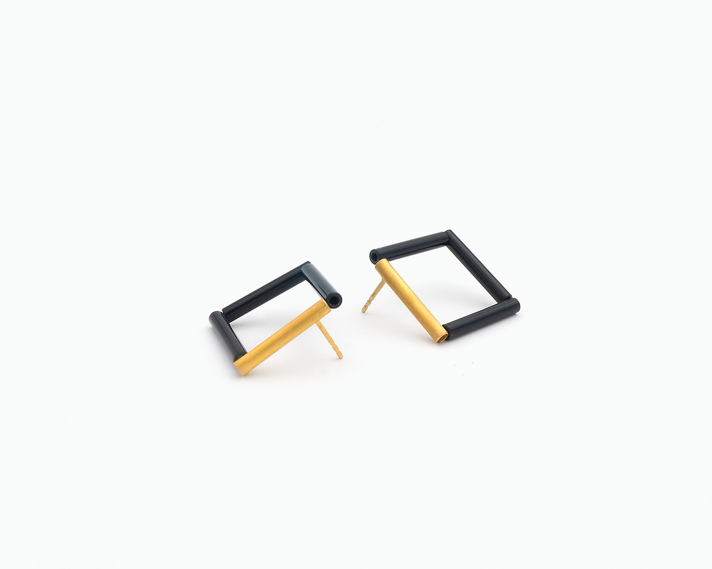 Zu sehen ist hier ein liegendes Ohrschmuck-Paar. Beide Ohrstecker sind Quadrate, kombiniert aus jeweils drei schwarzen Kunststoffröhrchen und einem silbernen, Feingold plattierten Röhrchen. Sie passen spiegelgleich zusammen. Sie liegen mit der Vorderseite nach oben zeigend versetzt zueinander und die hinten sitzenden Steckerstifte zeigen jeweils nach unten.