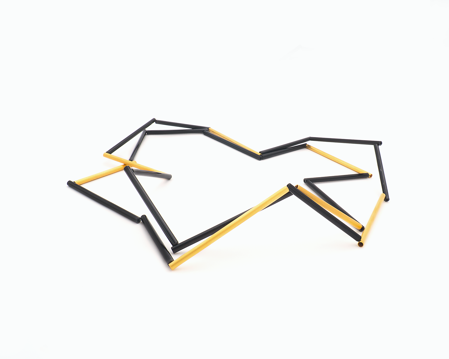 Zu sehen ist hier eine liegende Kette mit schwarzen und goldfarbenen Stäben. Die Stäbe sind Rohrabschnitte in zwei verschiedenen Längen. Die Kette ist doppelt gelegt dargestellt und stellt somit eine kurze Tragevariante dar. Die Kette hat eine grafische Anmutung, bei der die Linie als Hauptakteur dominiert.