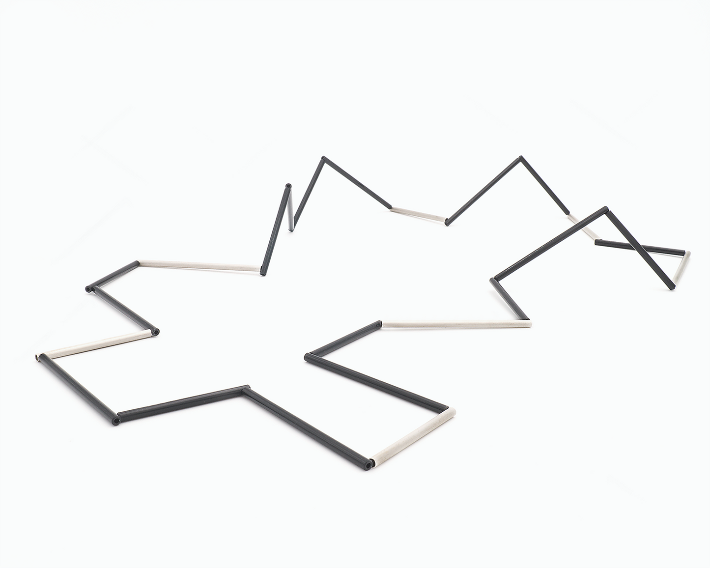 Zu sehen ist hier eine liegende Kette mit schwarzen und silbernen Stäben. Die Stäbe sind Rohrabschnitte in zwei verschiedenen Längen. Die Kette hat eine grafische Anmutung, bei der die Linie als Hauptakteur dominiert.