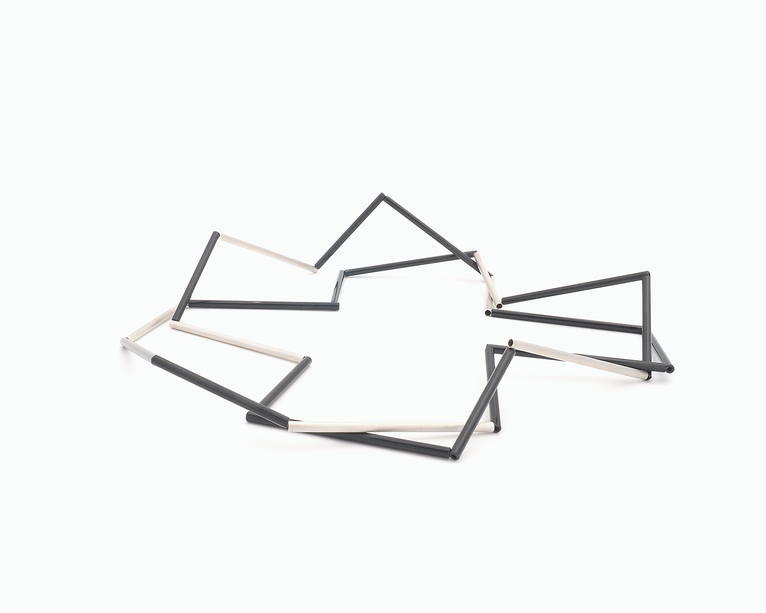 Zu sehen ist hier eine liegende Kette mit schwarzen und silbernen Stäben. Die Stäbe sind Rohrabschnitte in zwei verschiedenen Längen. Die Kette ist doppelt gelegt dargestellt und stellt somit eine kurze Tragevariante dar. Die Kette hat eine grafische Anmutung, bei der die Linie als Hauptakteur dominiert.