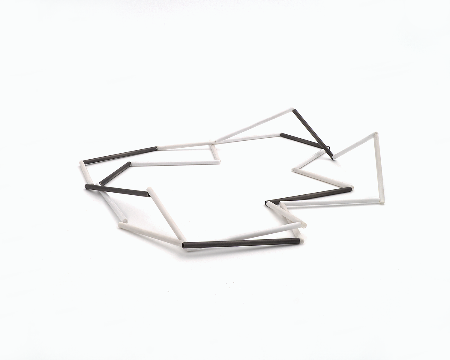 Zu sehen ist hier eine liegende Kette mit weißen und schwarzen Stäben. Die Stäbe sind Rohrabschnitte in zwei verschiedenen Längen. Die Kette ist doppelt gelegt dargestellt und stellt somit eine kurze Tragevariante dar. Die Kette hat eine grafische Anmutung, bei der die Linie als Hauptakteur dominiert.