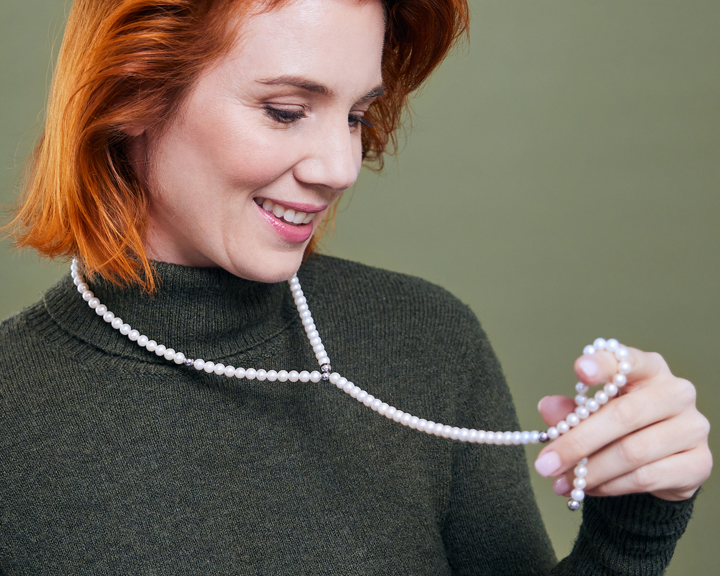 Zu sehen ist eine lächelnde Frau, die einen dunkelgrünen Rollkragenpullover trägt und darauf eine „chainge“-Kette mit weißen Süßwasserperlen. Ein Kettenende der Kette ist an eine Edelstahlkugel im Halsbereich angedockt. Die Frau hält den restlichen Kettenstrang in der Hand und betrachtet ihn.